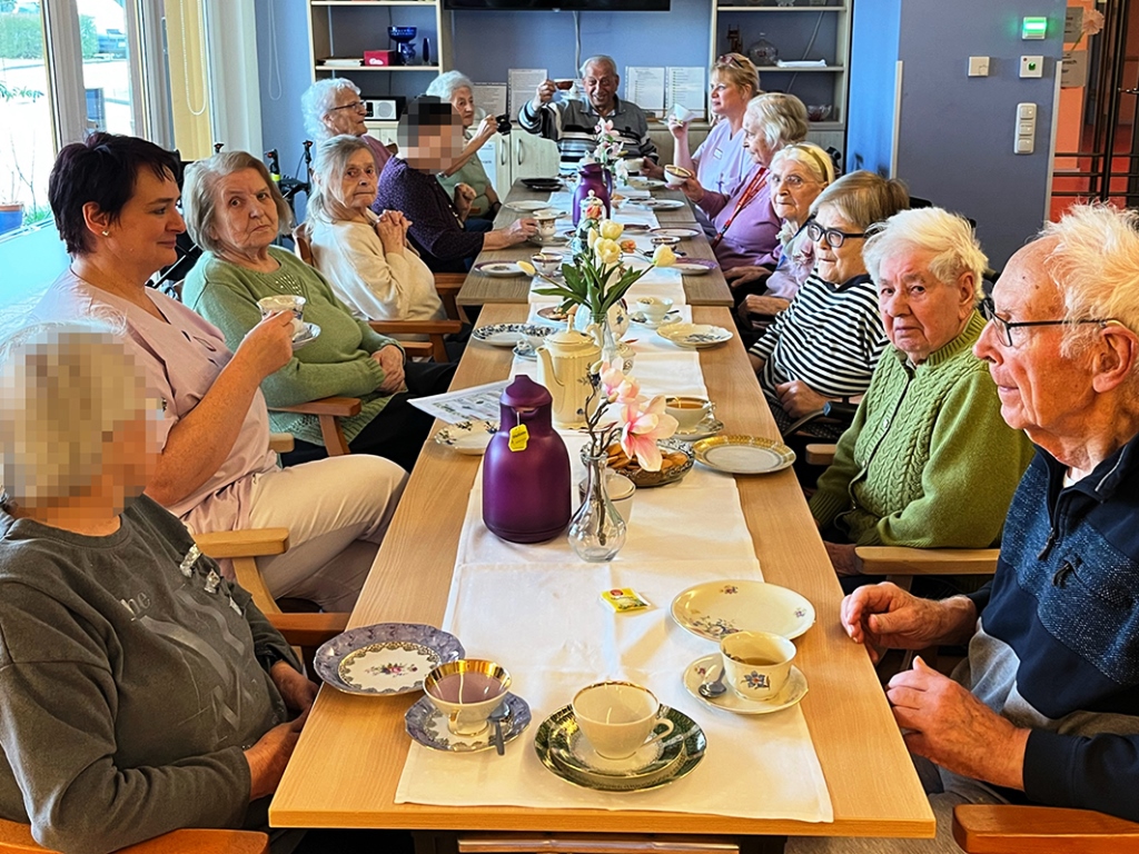 Eine Gruppe von Senioren und Pflegepersonal sitzt an einem langen Tisch und trinkt Tee.