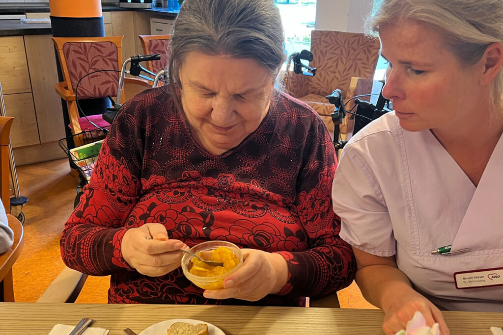 Eine &auml;ltere Frau, die ein kleines Glas mit Obst isst, sitzt neben einer Pflegekraft in einem Gemeinschaftsraum.