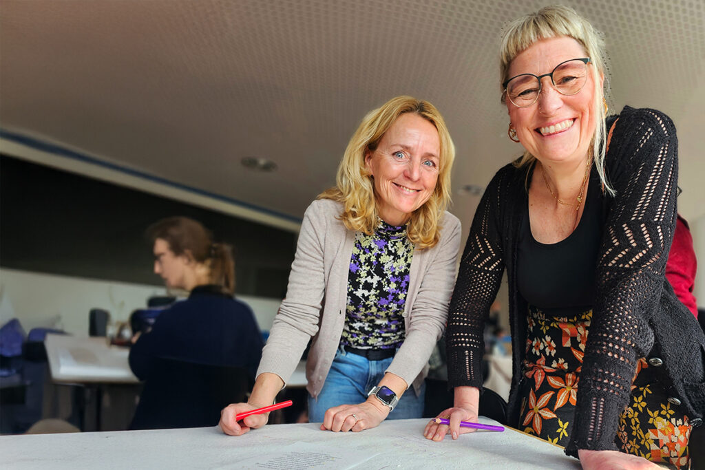 Zwei Frauen lehnen sich &uuml;ber einen Tisch und l&auml;cheln in die Kamera, w&auml;hrend sie bunte Stifte in den H&auml;nden halten.