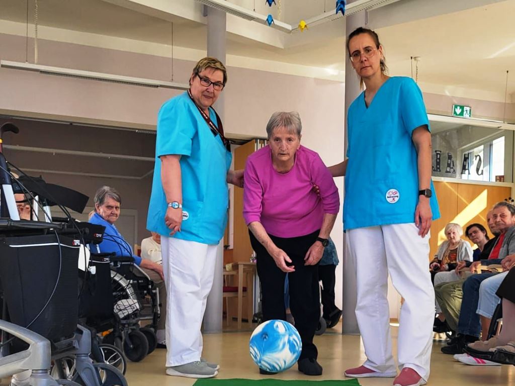 Eine ältere Frau in einem pinken Shirt steht mit Unterstützung von zwei Pflegekräften in einem Raum mit anderen sitzenden Personen.