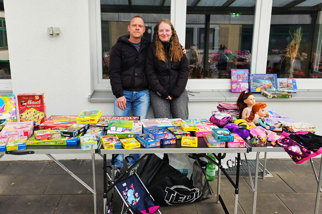Zwei Personen stehen hinter einem Tisch mit Spielsachen und Brettspielen im Freien.