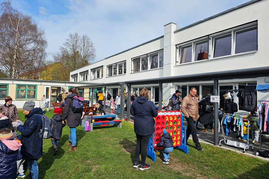Menschen versammeln sich im Freien vor einem modernen Geb&auml;ude mit gro&szlig;en Fenstern. Kinder spielen auf der Wiese und Erwachsene unterhalten sich. Ein gro&szlig;es Steckspiel ist zu sehen und Stand mit Kleidung im Hintergrund.