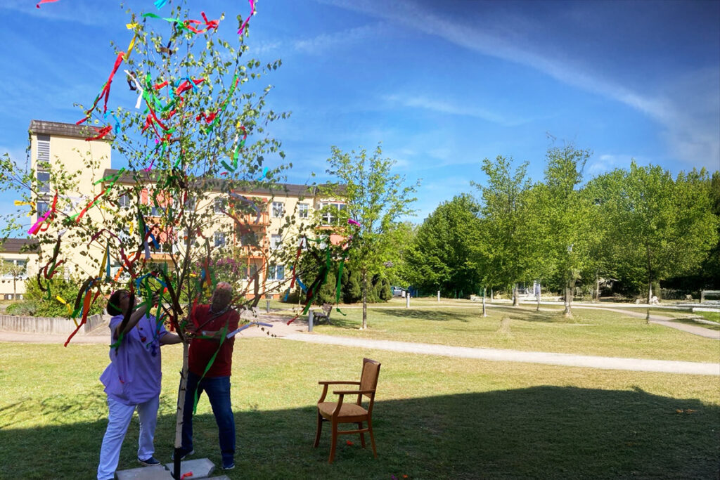 Zwei Personen schm&uuml;cken einen Baum mit bunten B&auml;ndern im Freien.