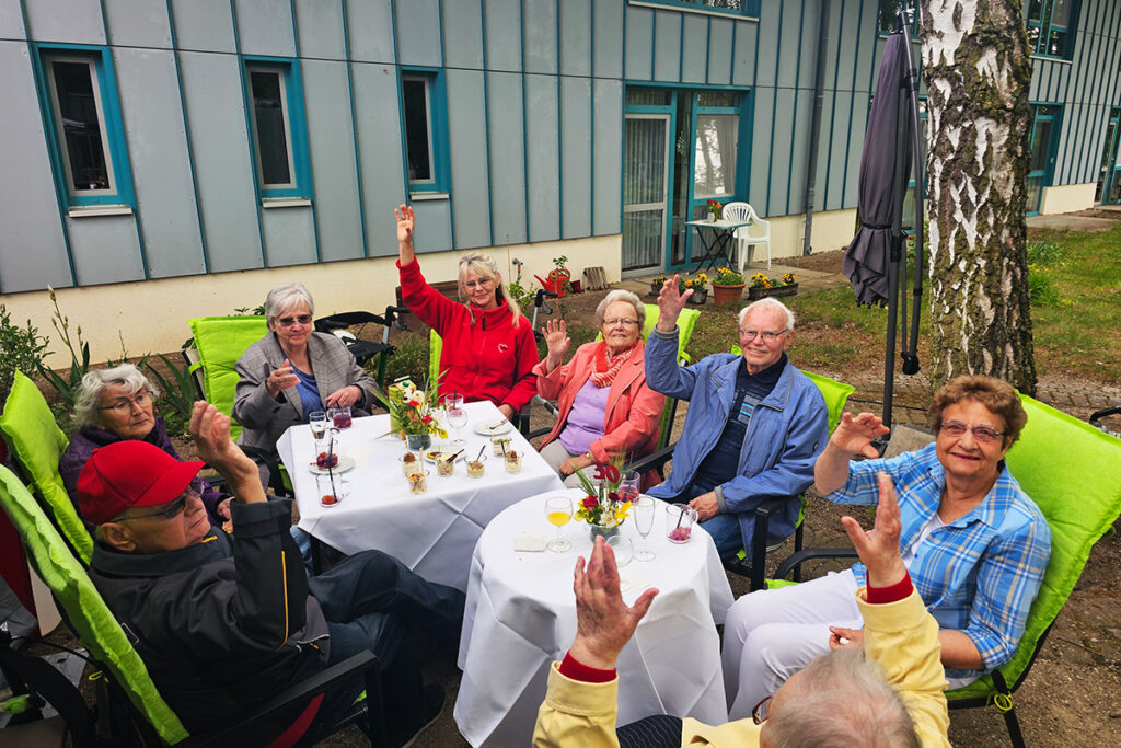 Gruppe von sieben &auml;lteren Menschen sitzt drau&szlig;en an einem Tisch und hebt die H&auml;nde.