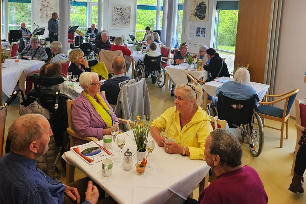 Eine Gruppe &auml;lterer Menschen sitzt an Tischen in einem Gemeinschaftsraum. Einige sind in Rollst&uuml;hlen, w&auml;hrend andere sich miteinander unterhalten. Im Hintergrund spielt eine Band.