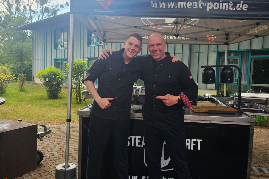 Zwei Personen in schwarzen Kochjacken stehen vor einem Grillstand und l&auml;cheln.