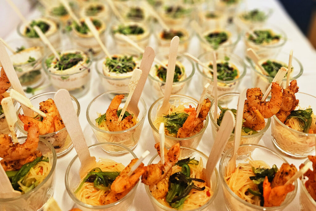 Mehrere kleine Gl&auml;ser mit Garnelenspie&szlig;en und Nudeln auf einem Tisch.