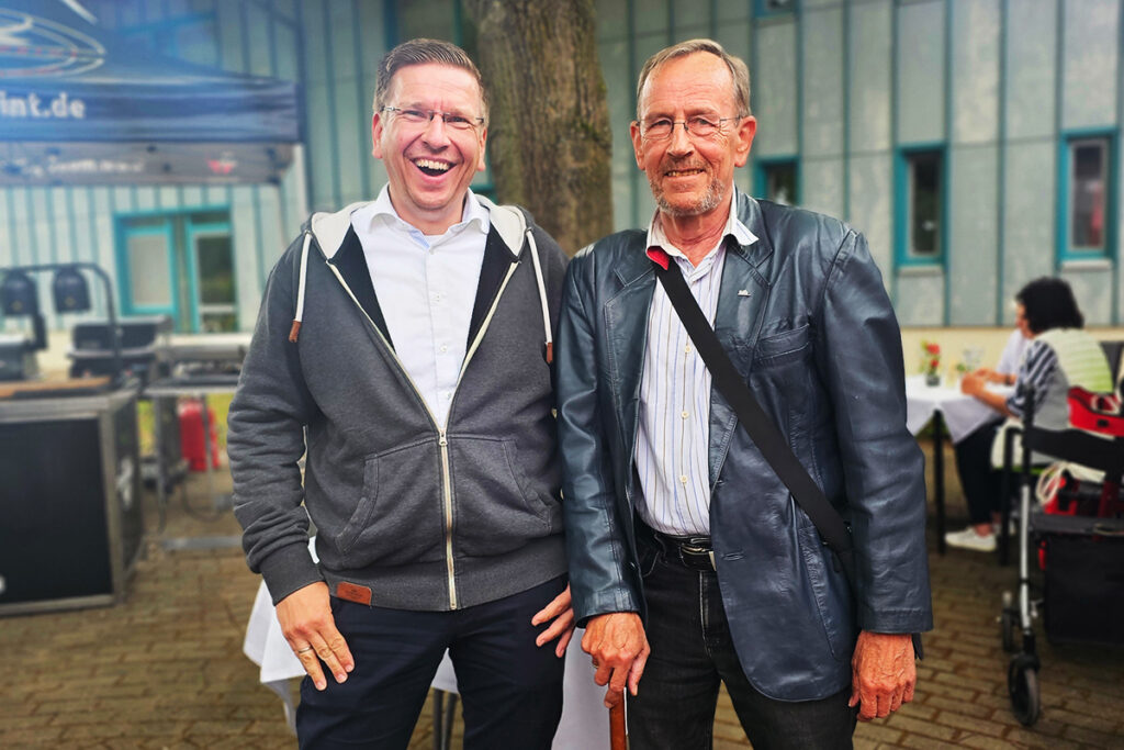 Zwei M&auml;nner stehen drau&szlig;en nebeneinander und l&auml;cheln in die Kamera.