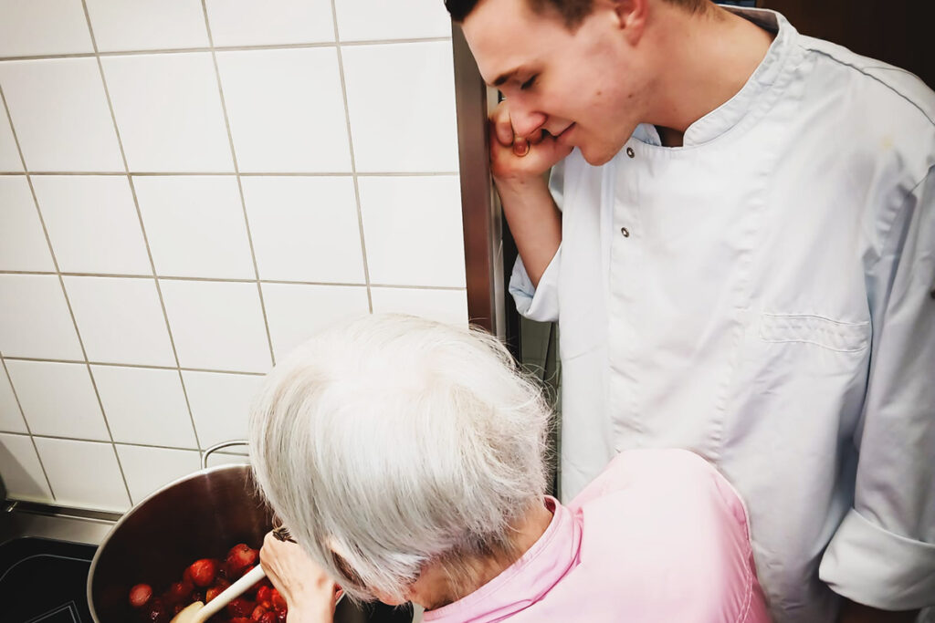 Eine ältere Person und eine jüngere Person in einer Küche, die zusammen an einem Topf arbeiten.