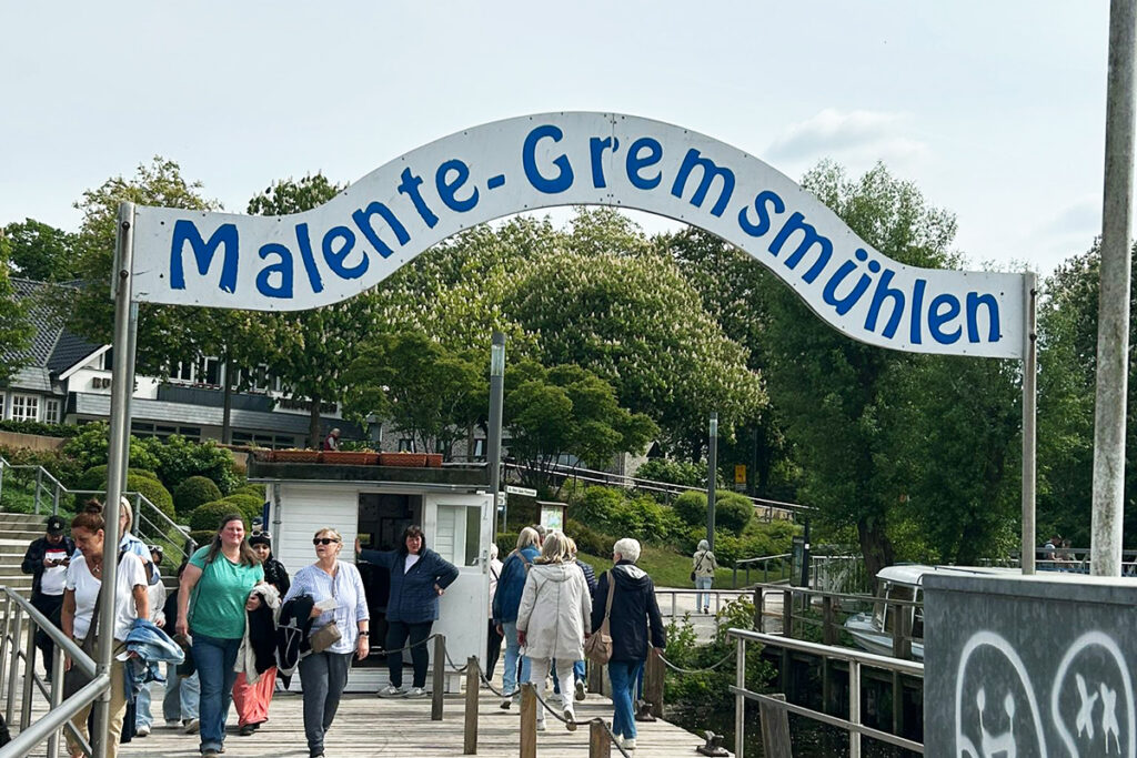 Ein Eingangsschild mit der Aufschrift 'Malente-Gremsm&uuml;hlen' &uuml;ber einem Steg, auf dem Menschen gehen.