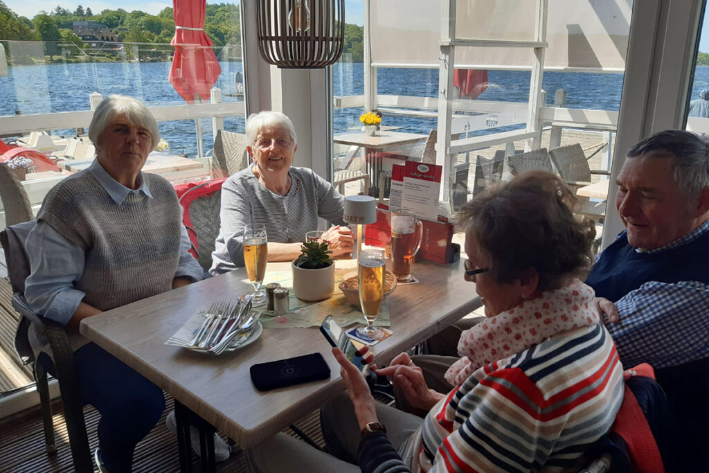 Eine Gruppe &auml;lterer Personen sitzt an einem Tisch in einem Caf&eacute; mit Blick auf einen See.
