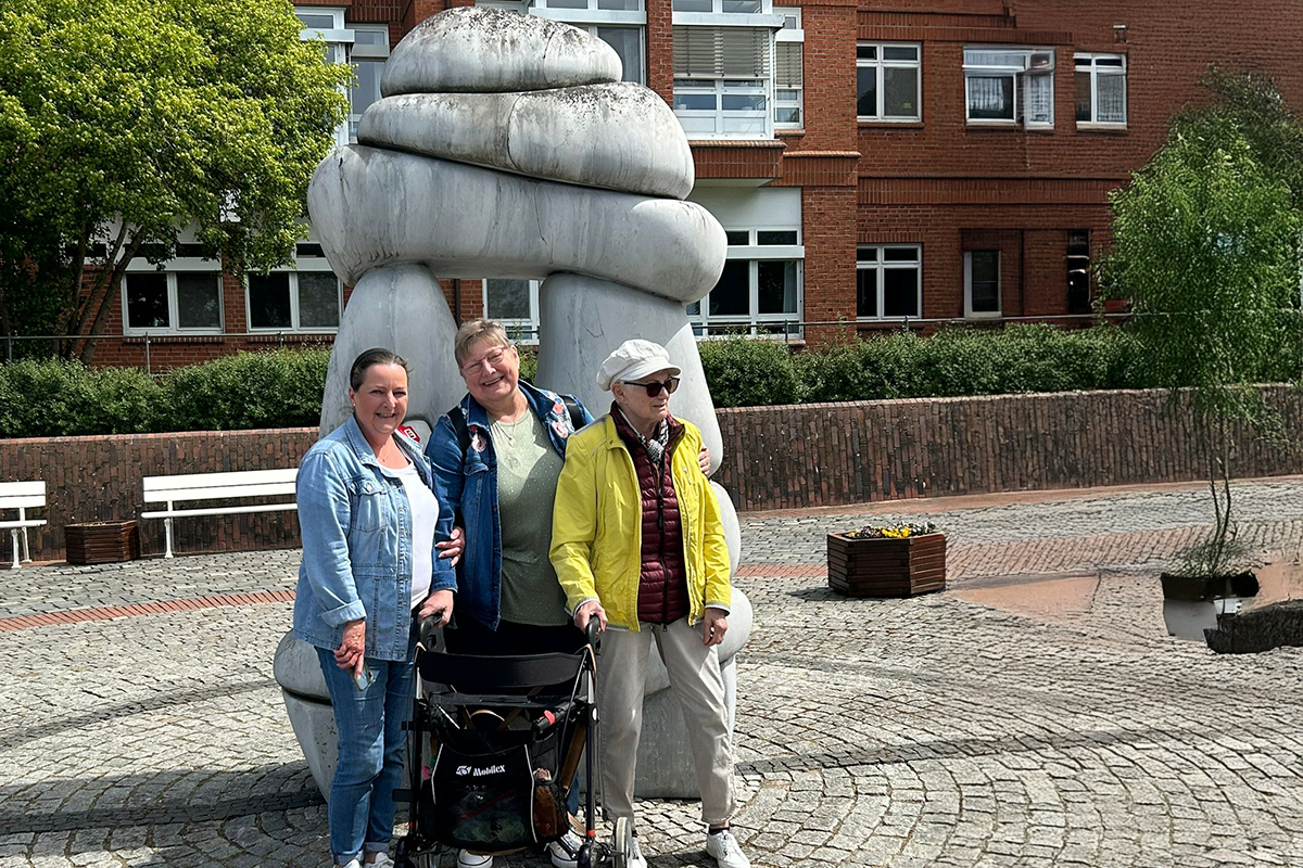 Drei Personen stehen vor einer großen weißen Skulptur in einer gepflasterten Außenanlage.