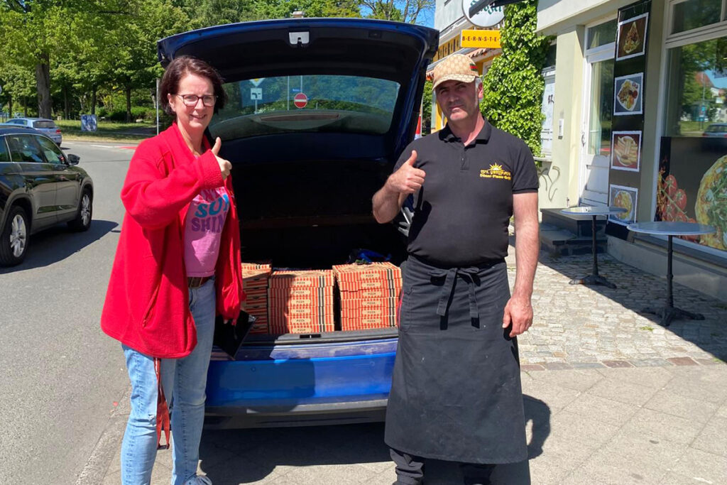 Eine Frau und ein Mann stehen vor einem offenen Kofferraum mit gestapelten Pizzakartons.