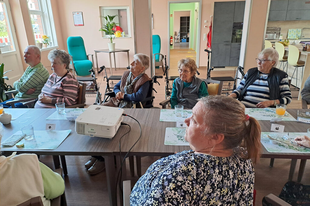 &Auml;ltere Menschen in einem Gemeinschaftsraum, die an einem Tisch sitzen und sich auf eine Pr&auml;sentation konzentrieren.