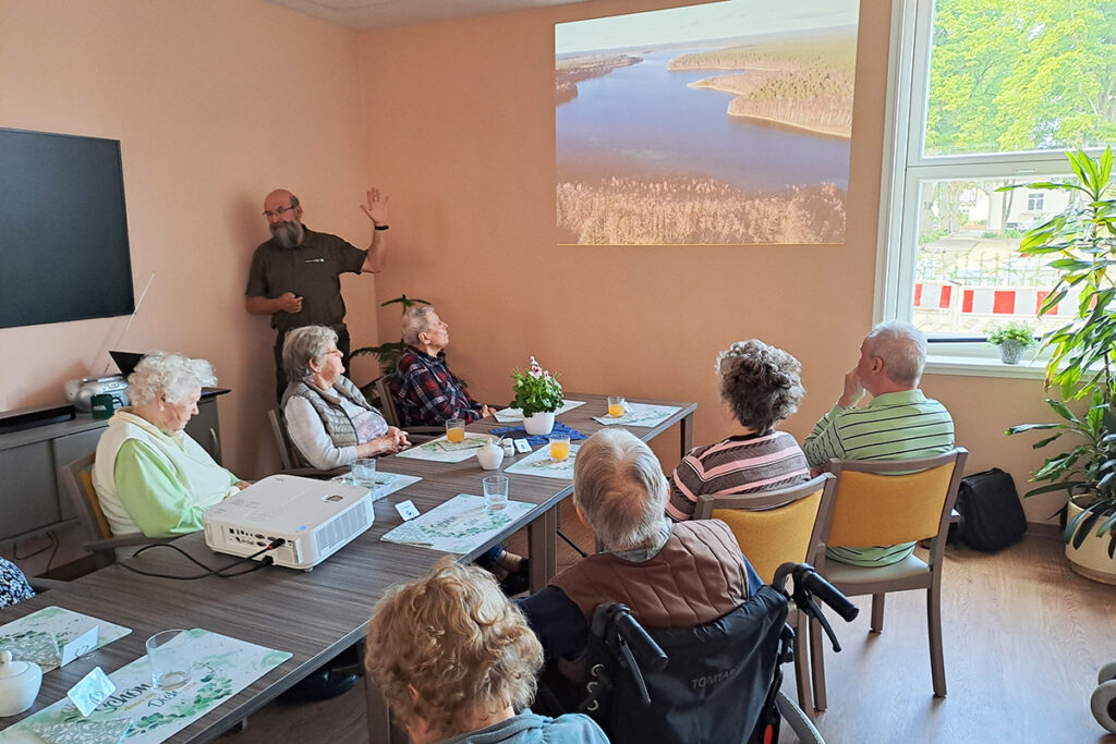 Ein Vortrag in einem Gemeinschaftsraum mit mehreren &auml;lteren Personen und einem Referenten.