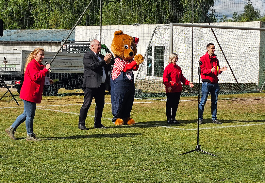 Gruppe von Menschen bei einer Veranstaltung auf einem Sportplatz, eine Person tr&auml;gt ein B&auml;renkost&uuml;m