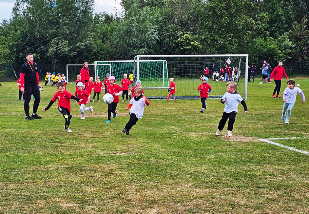 Kinder spielen Fu&szlig;ball auf einem Rasenplatz mit Erwachsenen als Betreuer.