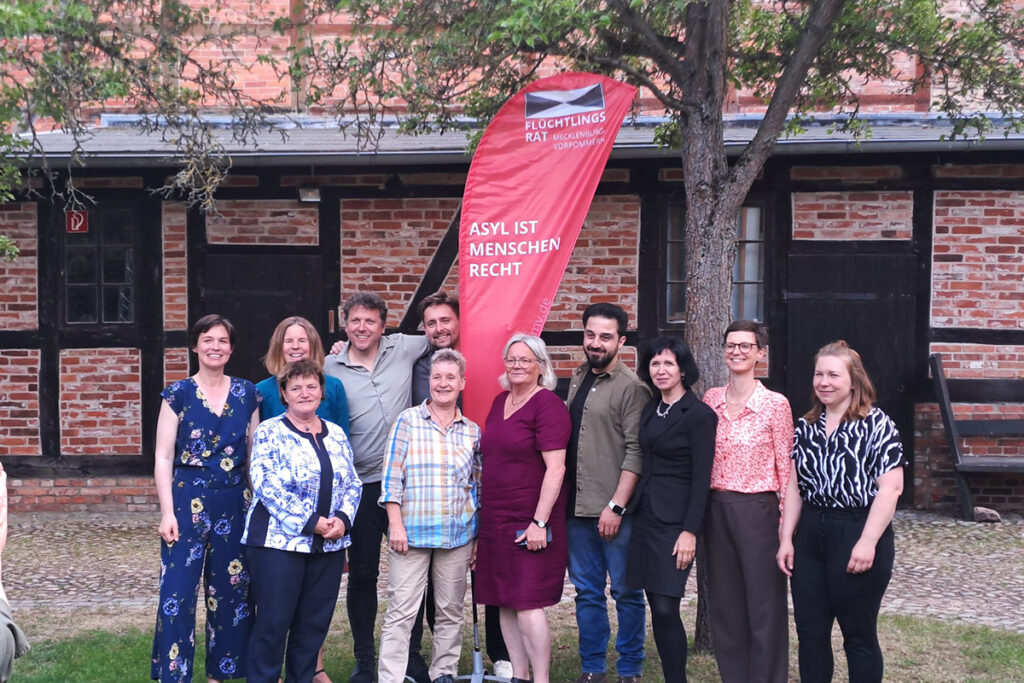 Eine Gruppe von Menschen steht drau&szlig;en vor einem Geb&auml;ude mit einem Banner, auf dem 'Asyl ist Menschenrecht' steht.