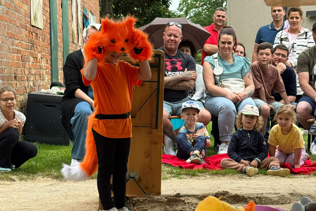 Ein Kind in einem orangefarbenen Fuchskost&uuml;m steht vor einem Publikum im Freien.