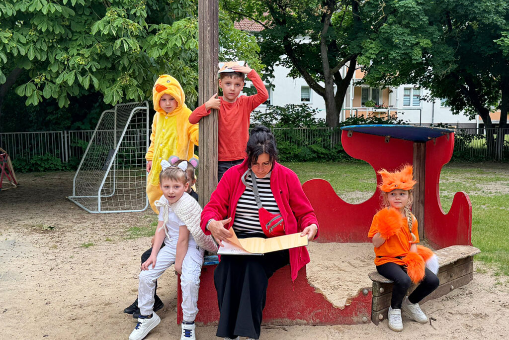 Frau liest einer Gruppe verkleideter Kinder auf einem Spielplatz vor.