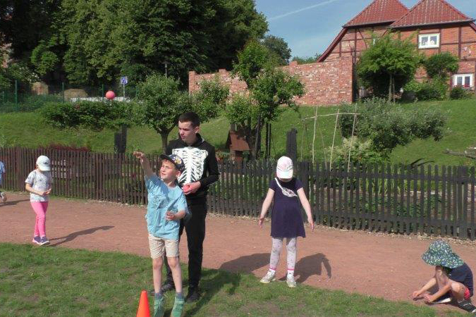 Ein Betreuer im Skelettkostüm hilft Kindern beim Ballspiel im Freien.