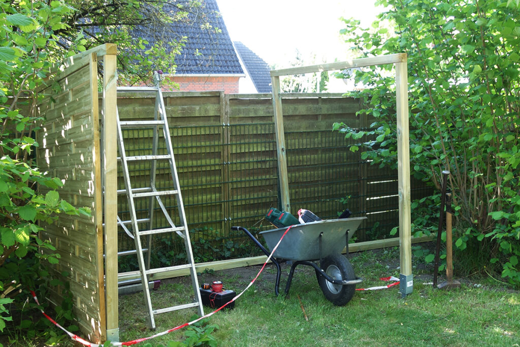 Eine teilweise errichtete Holzstruktur im Garten mit einer Leiter und einer Schubkarre, gef&uuml;llt mit Werkzeug.