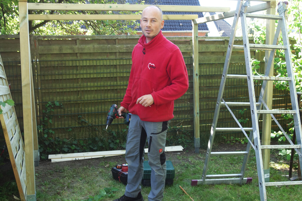 Ein Mann in rotem Pullover mit AWO-Logo arbeitet drau&szlig;en mit einer Bohrmaschine.
