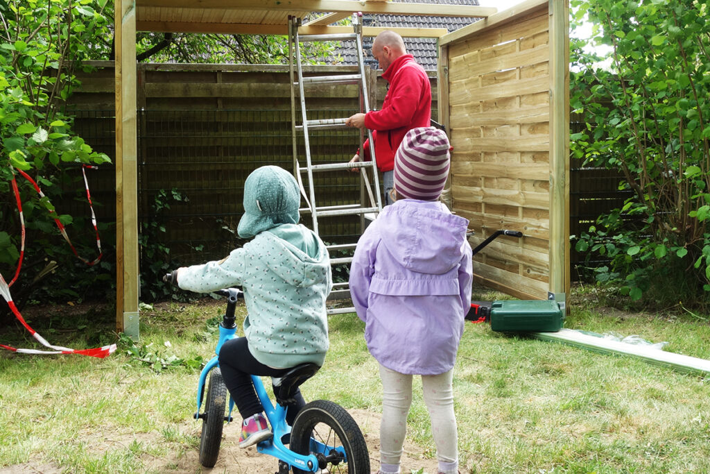 Ein Erwachsener in rotem Pullover arbeitet an einer Leiter, w&auml;hrend zwei Kinder zuschauen. Eines der Kinder sitzt auf einem kleinen Fahrrad, das andere steht daneben auf einer Wiese vor einem Holzunterstand.