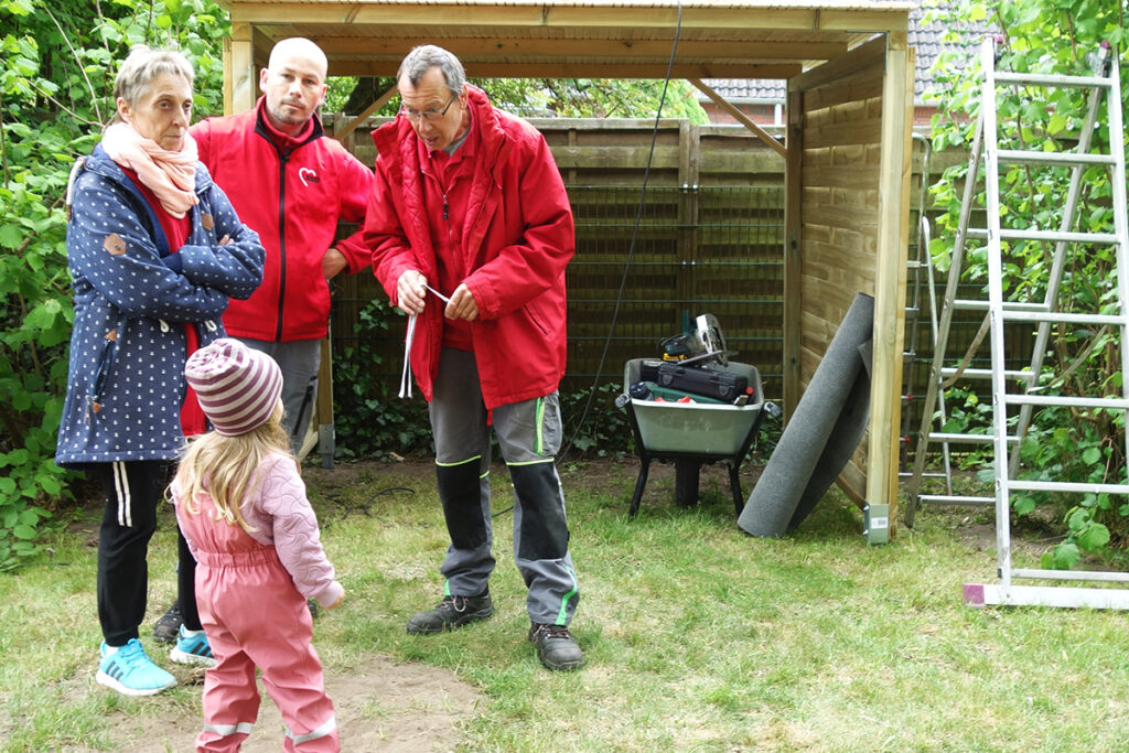 Vier Erwachsene und ein Kind stehen im Garten vor einem Holzunterstand.