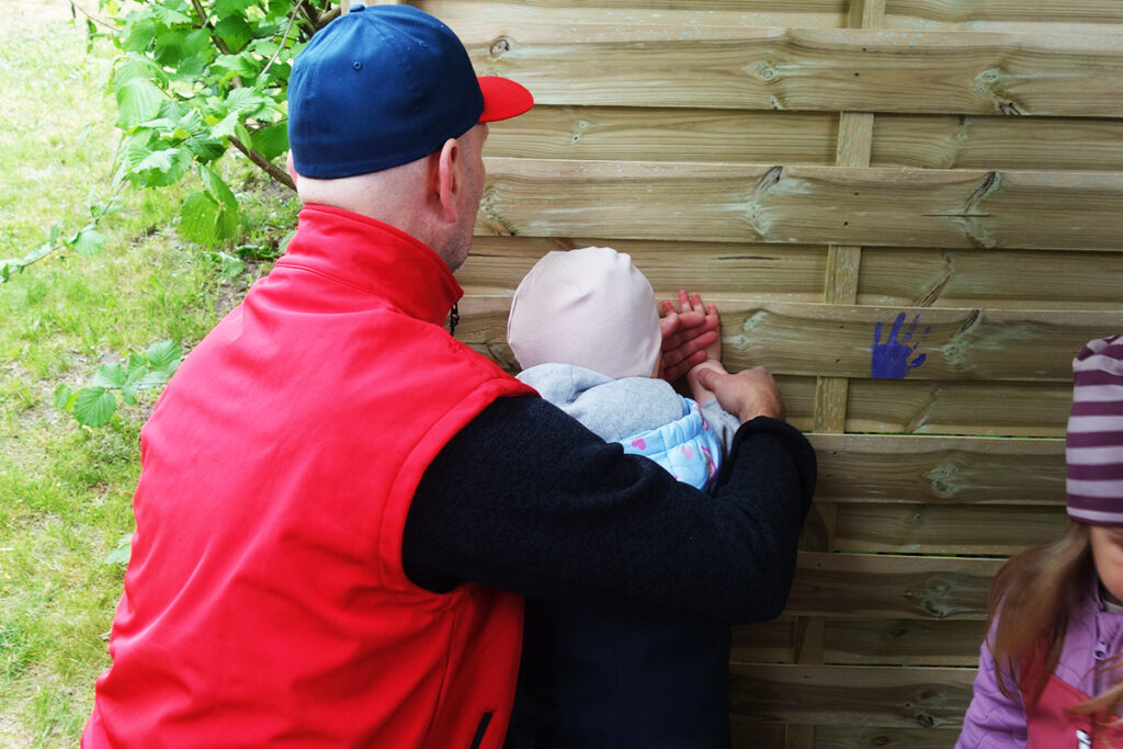 Ein Erwachsener mit roter Weste, der ein Kind an eine Holzwand f&uuml;hrt.