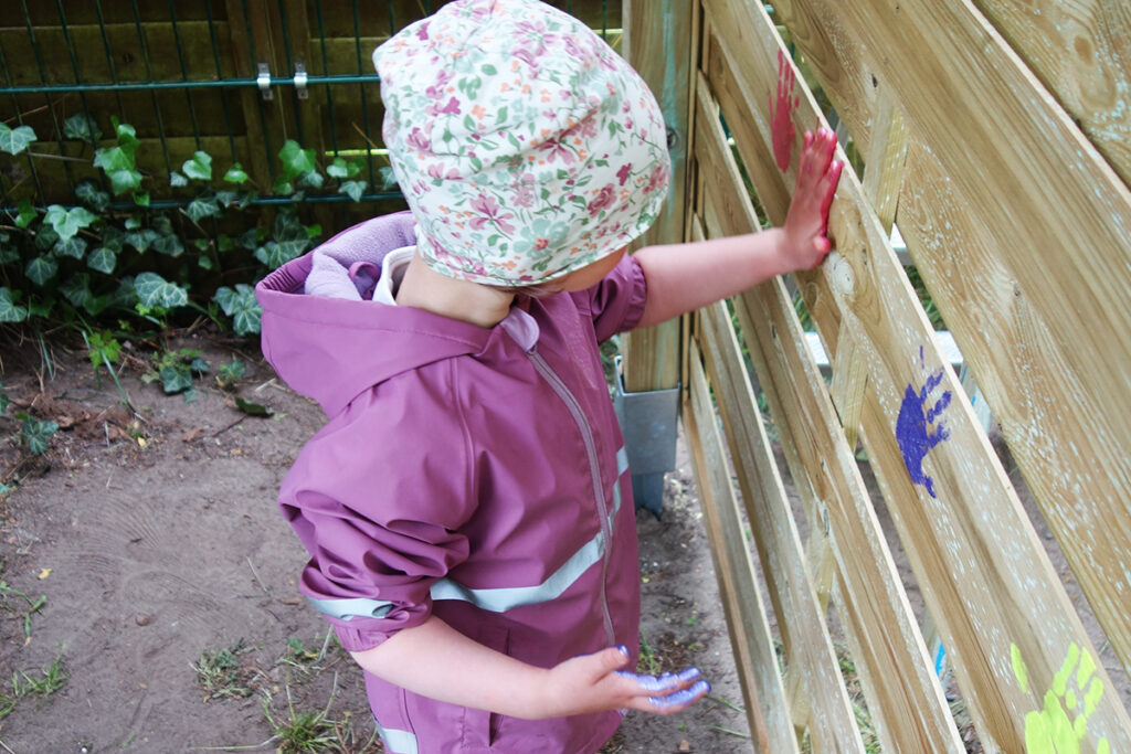 Ein kleines Kind in rosa Jacke malt Handabdr&uuml;cke auf einen Holzzaun.