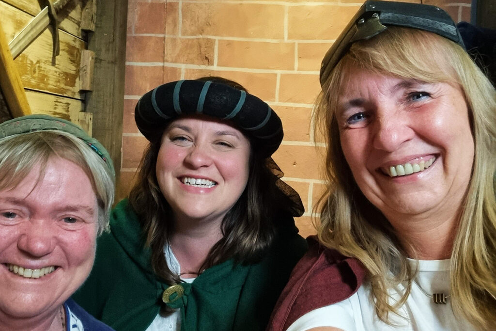 Drei l&auml;chelnde Personen in mittelalterlicher Kleidung vor einer Backsteinmauer