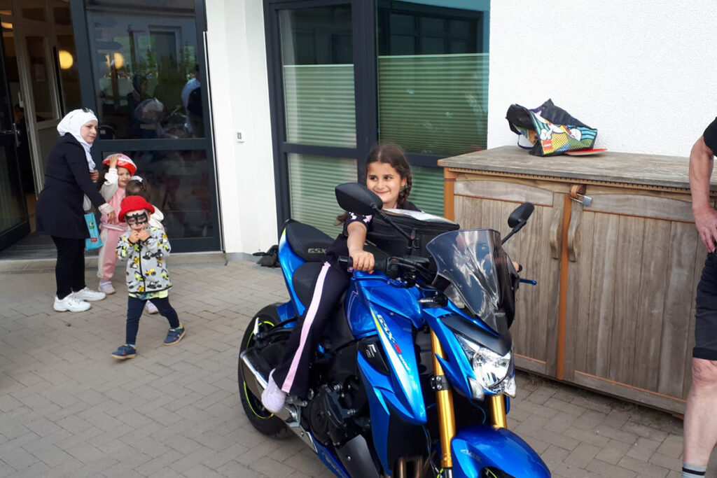 Ein M&auml;dchen sitzt auf einem blauen Motorrad vor einem Geb&auml;ude.