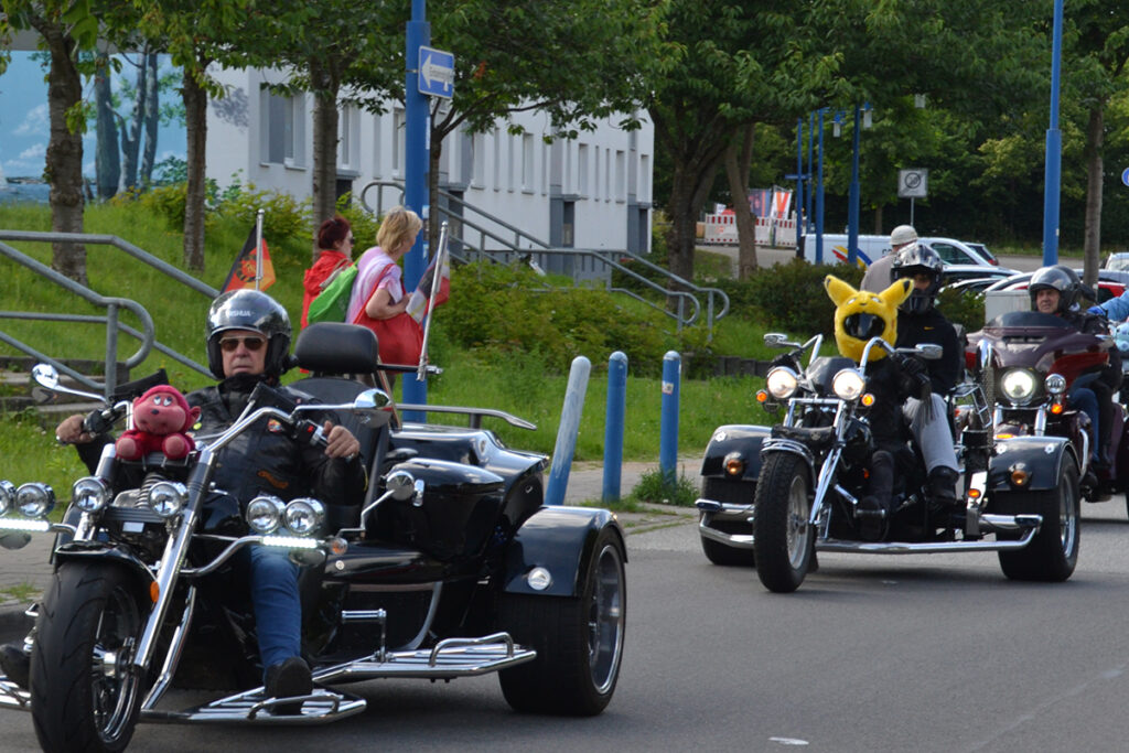 Eine Gruppe von Motorradfahrern auf dreir&auml;drigen Motorr&auml;dern f&auml;hrt eine Stra&szlig;e entlang. Der vordere Fahrer tr&auml;gt einen Helm und eine Jacke, w&auml;hrend vorne am Motorrad ein rotes Pl&uuml;schtier befestigt ist. Ein weiterer Fahrer hinter ihm tr&auml;gt einen auff&auml;lligen gelben Helm in Form eines bekannten Comic-Charakters. Im Hintergrund sind Passanten zu sehen.