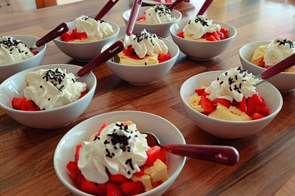 Mehrere Schalen mit einem Dessert aus Eiscreme, Erdbeeren, Schlagsahne und Schokoladenstreuseln, auf einem Tisch arrangiert