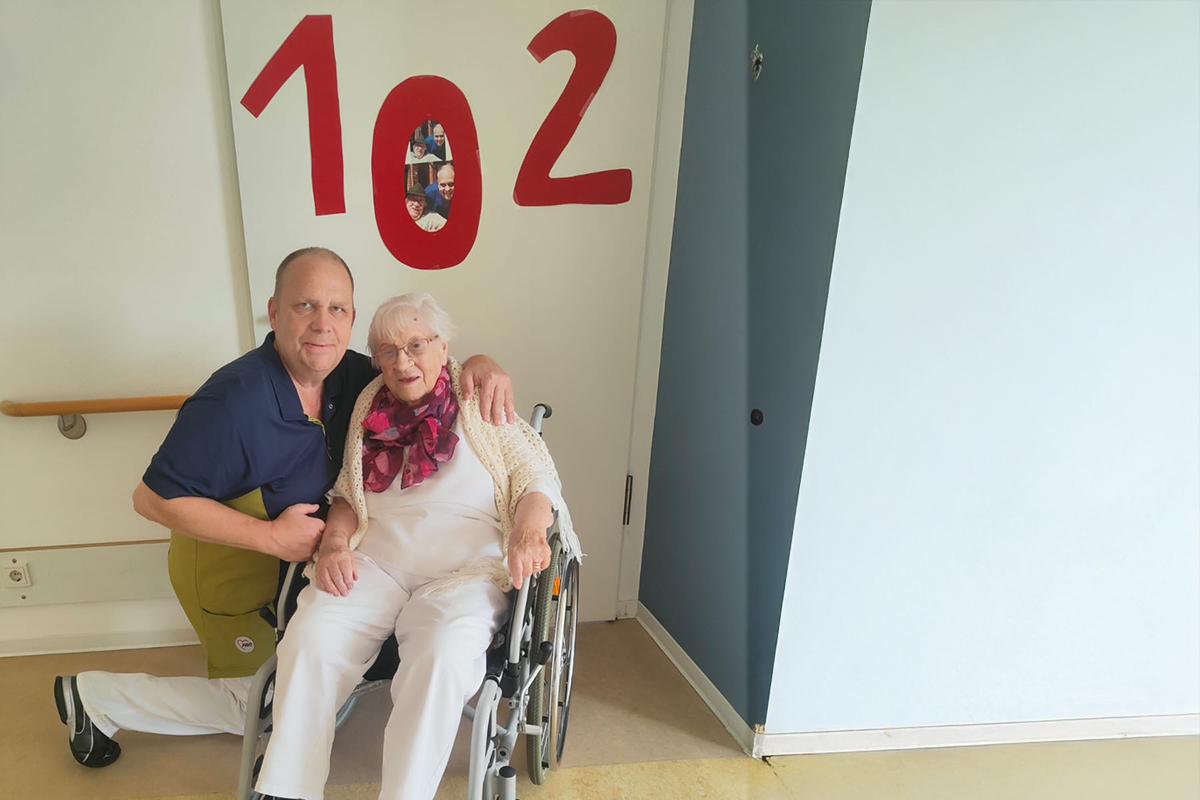 Eine ältere Frau im Rollstuhl neben einem Mann, der sie umarmt, vor einer Tür mit der Nummer 102.