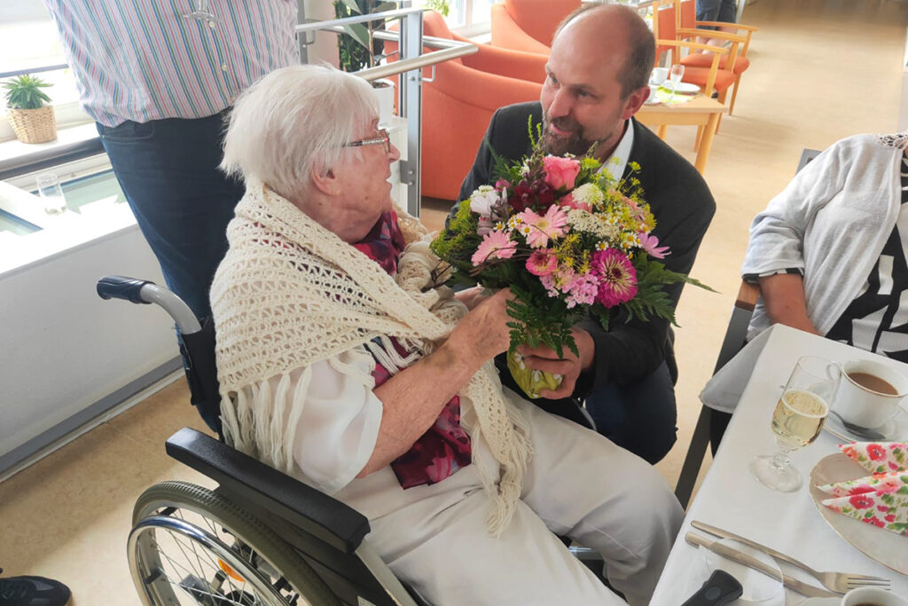 Eine &auml;ltere Frau im Rollstuhl bekommt einen Blumenstrau&szlig; von einem Mann &uuml;berreicht.