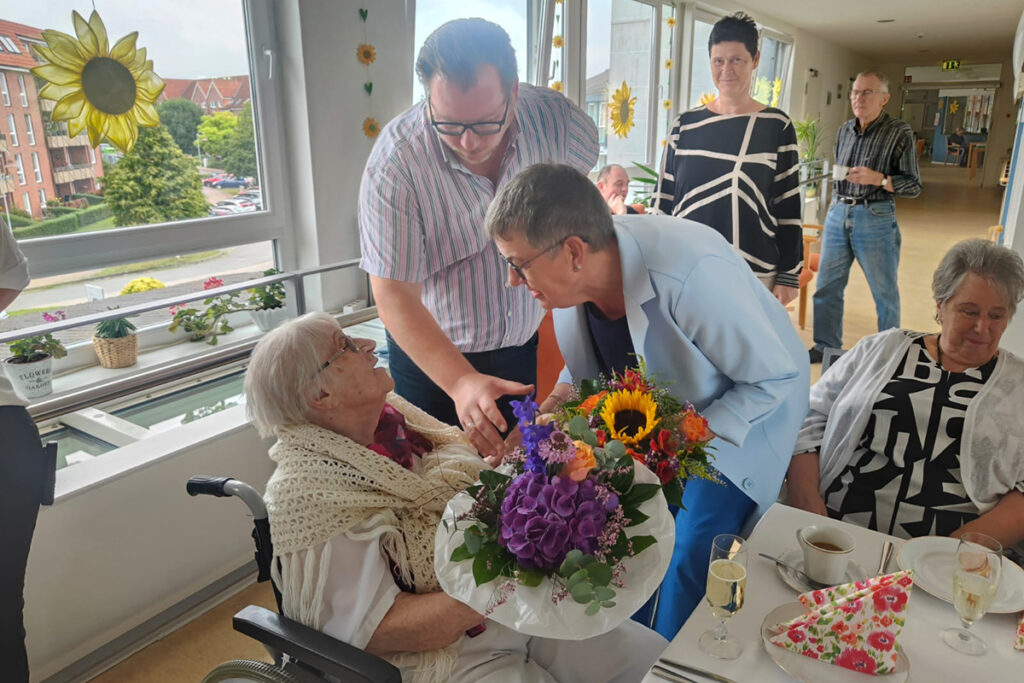 Person in Rollstuhl wird von Besuchern mit Blumenstrau&szlig; begr&uuml;&szlig;t