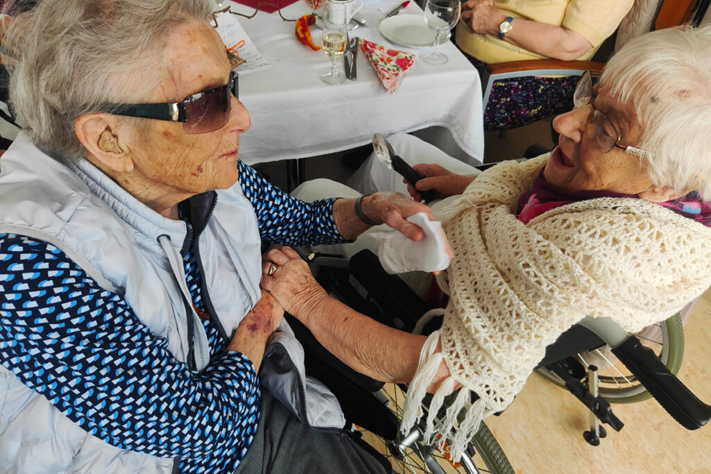 Zwei &auml;ltere Frauen unterhalten sich und l&auml;cheln einander an. Eine tr&auml;gt eine Sonnenbrille und einen Rollstuhl, die andere eine H&auml;kelstola.