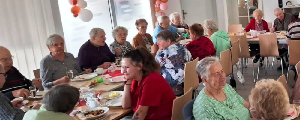 Eine Gruppe älterer Menschen bei einem gemeinsamen Essen in einem Raum, der mit Ballons geschmückt ist.