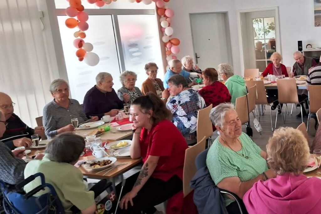 Eine Gruppe älterer Menschen bei einem gemeinsamen Essen in einem Raum, der mit Ballons geschmückt ist.
