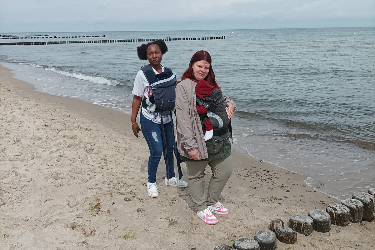Zwei Frauen mit Babys in Tragetüchern am Strand.