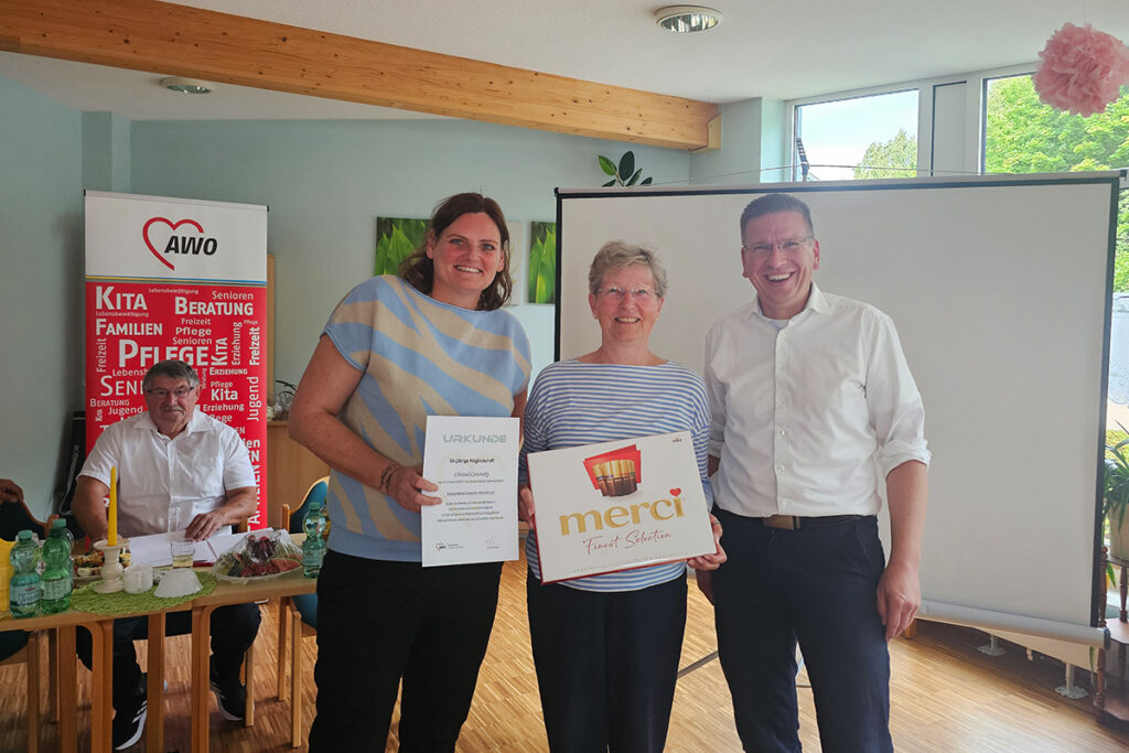 Ein Gruppenfoto von drei Personen, die bei einer AWO-Veranstaltung eine Urkunde und ein Merci-Schokoladenpaket pr&auml;sentieren.