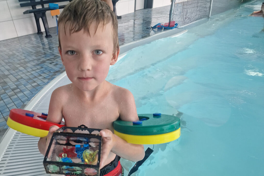 Ein Kind steht in einem Schwimmbecken, h&auml;lt eine kleine Box voller bunter Spielsachen und tr&auml;gt Schwimmfl&uuml;gel.