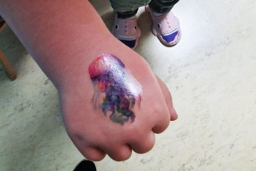 Eine Hand mit einem bunten Farbklecks und F&uuml;&szlig;e in pinken Hausschuhen