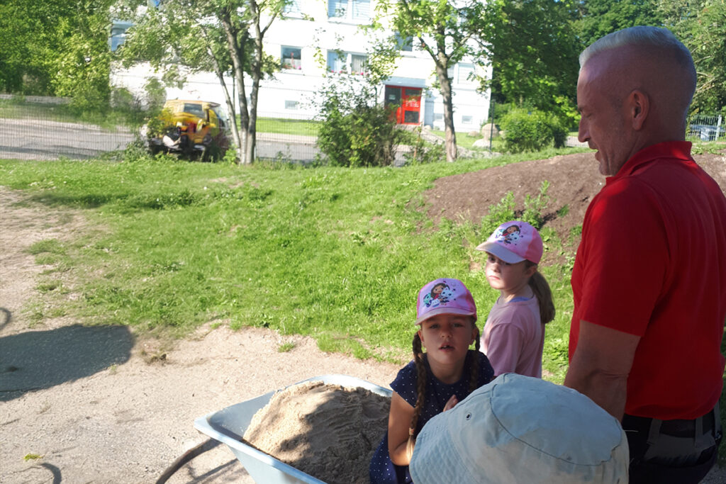Zwei Kinder mit Baseballkappen und ein Erwachsener stehen neben einer Schubkarre mit Sand in einem Garten.