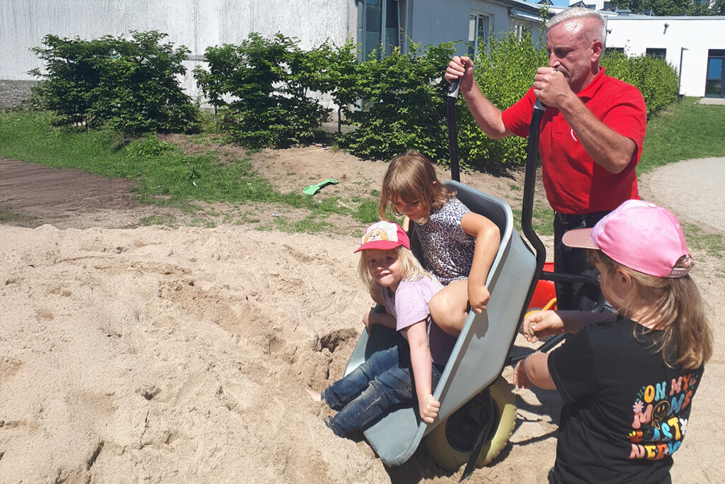 Ein Mann schiebt eine Schubkarre mit zwei Kindern, die im Sand spielen.