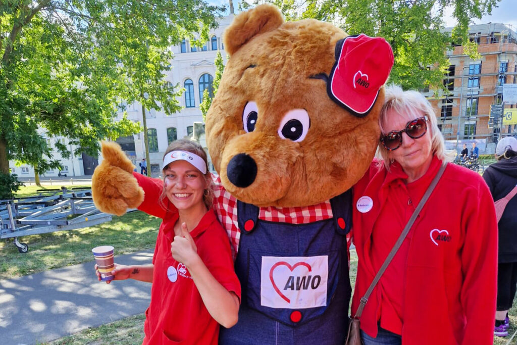 Drei Personen, darunter ein gro&szlig;es Maskottchen in B&auml;renkost&uuml;m, bei einer Veranstaltung im Freien.