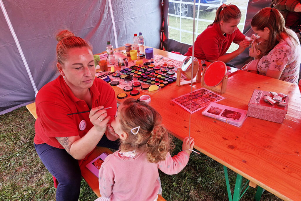 Zwei Erwachsene schminken zwei Kinder in einem Zelt mit Schminkutensilien auf einem Tisch