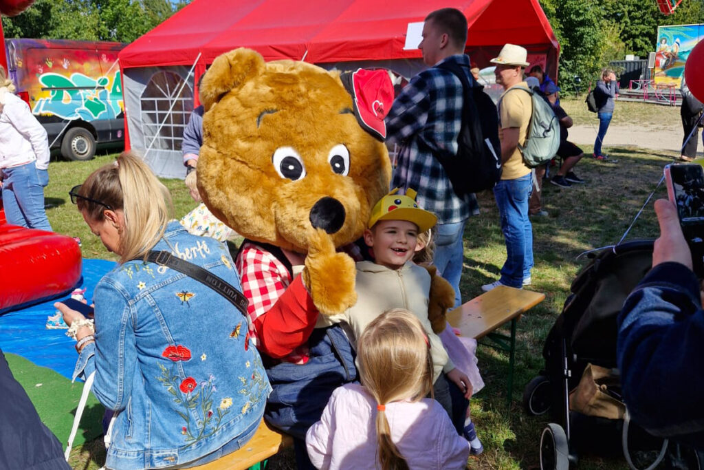 Szene von einer Veranstaltung im Freien mit einem gro&szlig;en Teddyb&auml;r-Kost&uuml;m und mehreren Personen, darunter Kinder